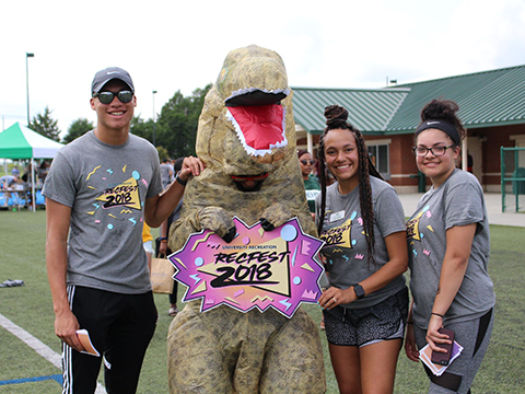 3 people standing next to a dinosaur holding a sign that reads RECFEST 2018.