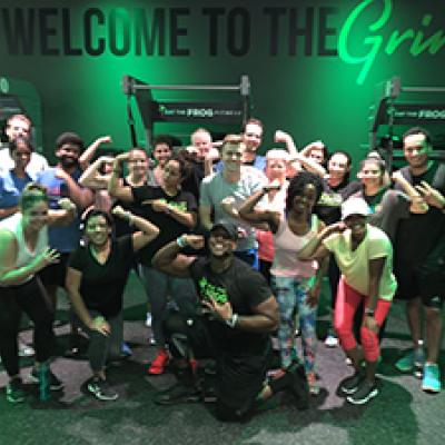 Group of individuals posing in a gym with the words "Welcome to the Grind" on the wall.